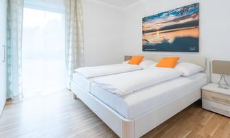A modern bedroom with a large double bed and orange cushions. A picture of a sunset hangs on the wall, and large windows provide plenty of daylight.