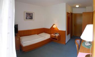 A simple single room with a bed, desk, and television. The walls are painted light, and there is a lamp as well as a window with a curtain.