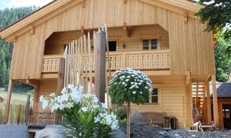 A cozy wooden house with a balcony and an entrance made of wooden planks. Blooming plants stand in front of the house, and it is surrounded by a green landscape.