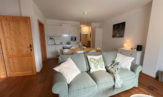 A bright and inviting living room with a green sofa and an open kitchen. The wooden details create a cozy atmosphere.