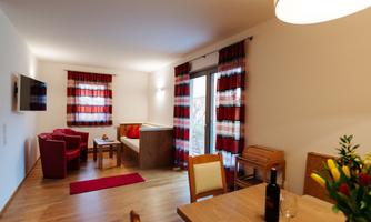 A cozy living space with red chairs and a wooden table. Large windows let in plenty of light, and the floor is made of wood.