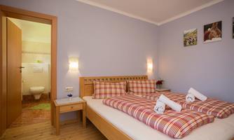 A bright bedroom with a double bed and checked pillows. In the background, a bathroom is visible.