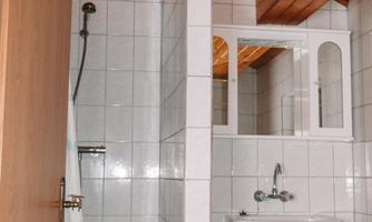 A simple bathroom with a shower and a sink. The walls are covered with white tiles and the ceiling has wooden cladding.