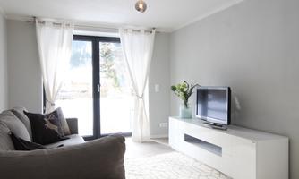A cozy living room with a gray sofa and a modern television. Large windows with curtains let in plenty of light.