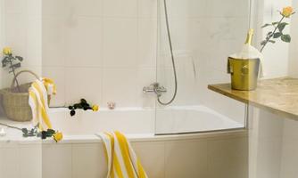 A modern bathroom with a bathtub and a shower. Yellow striped towels are lying on the floor, and there are decorative flowers on the shelf.