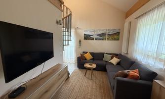 A cozy living room corner with a large TV, a gray sofa, and a small table. The walls are light, and there are pictures of landscapes as well as large windows that let in plenty of light.