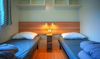 A simple room with two single beds and wooden flooring. The walls are light, and there is a shelf above the beds.