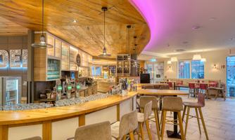 A modern restaurant with a wooden counter area and stylish bar stools. Soft lighting creates an inviting atmosphere.