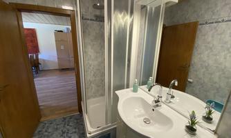 A modern bathroom with a shower and washbasin. In the background, a living area can be seen.