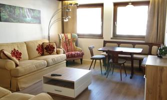 A cozy living room with beige sofas and decorative cushions. A table and chairs are also available, as well as large windows for plenty of daylight.