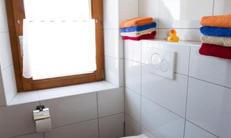 A modern bathroom with a white toilet and a window. Colorful towels are neatly stacked on a shelf.