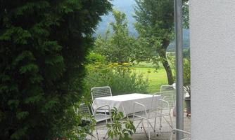 A quiet outdoor area with a table and chairs on a paved floor. In the background, green meadows and trees can be seen.