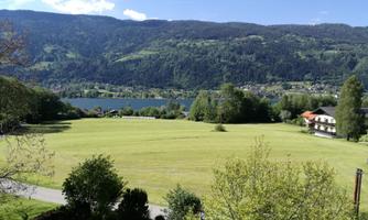A beautiful landscape with a vast green field and a clear lake. Gentle mountains rise in the background and a blue sky.