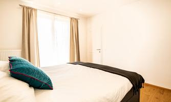 A bright bedroom with a large bed and decorative pillows. Bright curtains hang in front of the windows, letting in daylight.