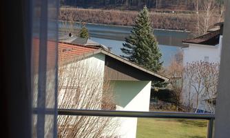 A balcony with a small table and chairs offers a view of a lake and surrounding mountains. The nature is calm and inviting.
