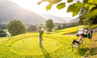 A golfer hits the ball on a picturesque golf round in a mountainous landscape. In the background, green hills and a golf cart can be seen.
