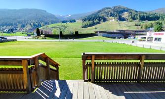 A view from a terrace overlooking a green meadow, surrounded by mountains. The sky is clear and sunny.