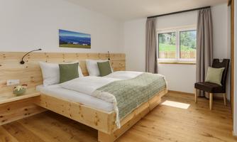 A modern bedroom with a wooden double bed. Large windows let in plenty of daylight and offer a view of the greenery.
