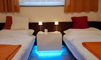 A cozy bedroom with two single beds, orange bedding, and modern nightstands. The blue lighting creates a relaxed atmosphere.