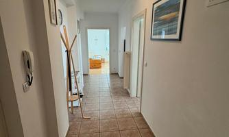 A bright hallway with tiled floor and white walls. At the end of the hallway, an open door is visible.