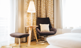 A cozy armchair with a cushion and a floor lamp, next to a small table. The room is bright and cheerful, with curtains and a warm wooden floor.