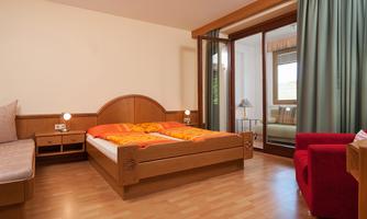 A cozy hotel room with a double bed and colorful bedspreads. There is a comfortable armchair and large windows that let in daylight.