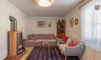 A cozy living room with a large sofa, a carpet, and a heating stove. Pictures hang on the wall, and a wooden decoration is visible.