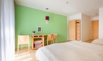 A modern room with green wall color and two single beds. A small table with chairs and a shelf complements the furnishings.