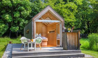A small, modern holiday home with wooden walls stands amidst green grass. In front of the terrace, there are two chairs and a table that looks inviting.