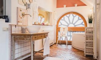 A bright bathroom with a large window and a round window frame. There is a wooden dresser with a sink and a cozy atmosphere.