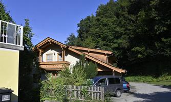 A cozy house with wooden siding, surrounded by trees. A car is parked on the gravel area in front of the house.