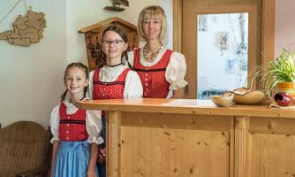 Three women in traditional attire are standing at a reception desk. In the background, wooden decorative elements are visible.