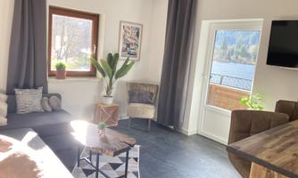A modern living room with a cozy couch and a coffee table. Large windows offer a beautiful view of the outdoor area with water and plants.
