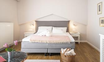A modern bedroom with a large double bed and soft color tones. Next to the bed is a small table and a laundry basket.