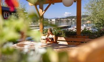 A cozy wooden terrace by the lake with a young woman relaxing and reading a book. The surroundings are green, with a view of the water and a clear sky.