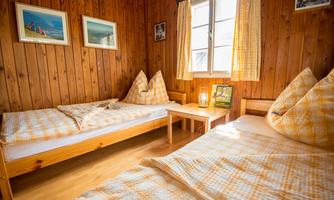 A cozy room with wooden paneling, two beds, and bright curtains. A small table sits between the beds, and pictures hang on the wall.