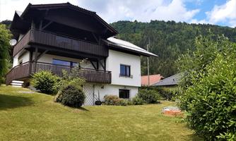A beautiful house in the greenery with a balcony and a well-kept lawn. In the background, there are mountains and a blue sky.