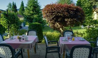 An inviting outdoor breakfast area with two tables covered in pink tablecloths. In the background, a well-kept garden with green trees and shrubs can be seen.