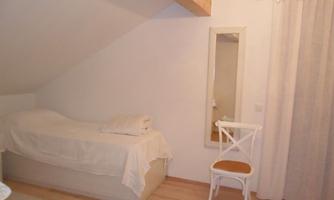 A cozy room with a bed, a chair, and a mirror. The walls are bright and the decor is simple and welcoming.