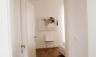 A bright room with a white chair and a painting on the wall. The floor is made of wood and the walls are simple and light.