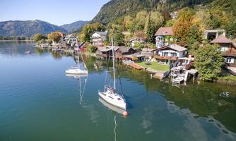 A tranquil lake landscape with sailboats and picturesque houses along the shore. Trees and mountains surround the scene, creating a peaceful atmosphere.