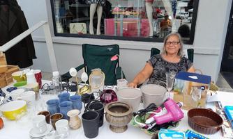 Eine ältere Frau sitzt an einem Tisch voller Haushaltswaren und Töpfen. Im Hintergrund sind ausgestellte Kleider und weitere Gegenstände zu sehen.