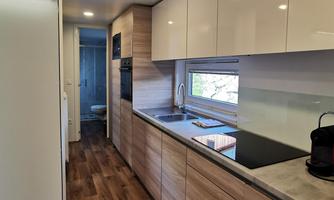 A modern kitchen with wooden shelves and bright surfaces. It features large windows that let in plenty of light.
