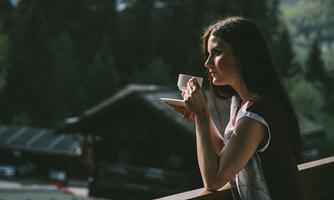 A woman is standing on a balcony holding a cup in her hand. In the background, trees and buildings can be seen.