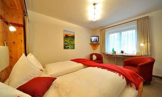 A cozy room with a double bed and red pillows. There is a television, windows with curtains, and a small seating area.