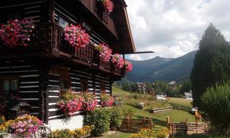 A traditional wooden house with blooming flowers on the balcony. In the background, green mountains and an idyllic landscape can be seen.