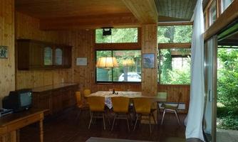 A cozy living space with wooden walls and large windows. The room is equipped with a dining table, chairs, and a television.