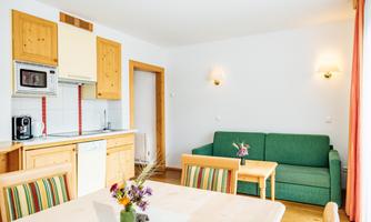 A modern living room with a green couch and a table. The kitchen is well-equipped with wooden cabinets and appliances.