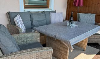 A cozy seating area on a terrace with a table and comfortable rattan furniture. In the background, there is a sunshade and a wooden cabinet.