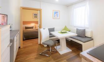 A modern room with a dining area and a cozy bed in the background. The walls are light, and there are large windows that let in plenty of natural light.